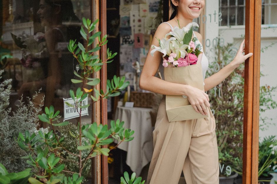 A smiling woman holding a beige paper bag filled with fresh flowers, including white lilies and pink roses, standing at the entrance of a flower shop with large glass windows. The bouquet showcases vi