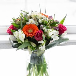 A masterfully composed bouquet, this floral creation reflects the community spirit and natural charm of Osidge. Cream-hued gerberas with plush petals form a luminous circle around glowing orange centres, recalling spirited gatherings or spring events at the nearby Osidge Lane green spaces. Deep magenta roses provide a sophisticated note, while clusters of white alstroemeria and petite filler flowers breathe brightness and texture into the arrangement. Blush pink accents are nestled for a playful touch, adding to the sense of layered abundance. The rich green foliage-anchored by large, curved aspidistra leaves-embraces the bouquet in a protective collar, echoing the tranquility and lushness found in Osidge's parks and leafy streets. Slender, pointed leaves and airy sprigs of greenery fill the interior, offering natural contrast and form. Arranged in a tall, pristine glass vase and illuminated by soft daylight, the bouquet's colours appear vibrant yet soothing. A gentle floral fragrance, mingled with garden-fresh notes, invites lingering appreciation. Whether as a centrepiece for a family celebration or a thoughtful gesture of thanks, this bouquet, artfully designed by the local team at Flowers Osidge, exudes joy, optimism, and heartfelt care-an ode to the connections that make Osidge special.
